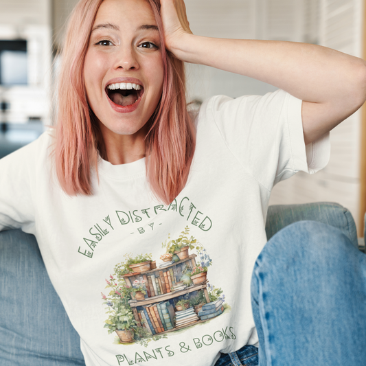 Woman shown wearing our Easily Distracted by Plants & Books T-Shirt Featuring a charming watercolor design of a books shelf filled with books and several potted plants on a natural light tan  colored t-shirt.